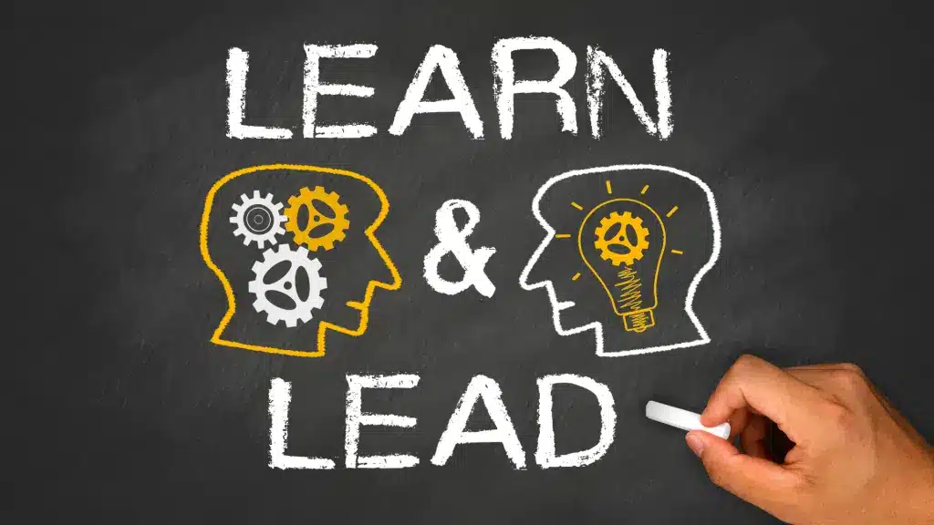 Chalkboard drawing showing two heads, one with gears and the other with a lightbulb, representing learning and leadership, with the text 'Learn & Lead' written above.