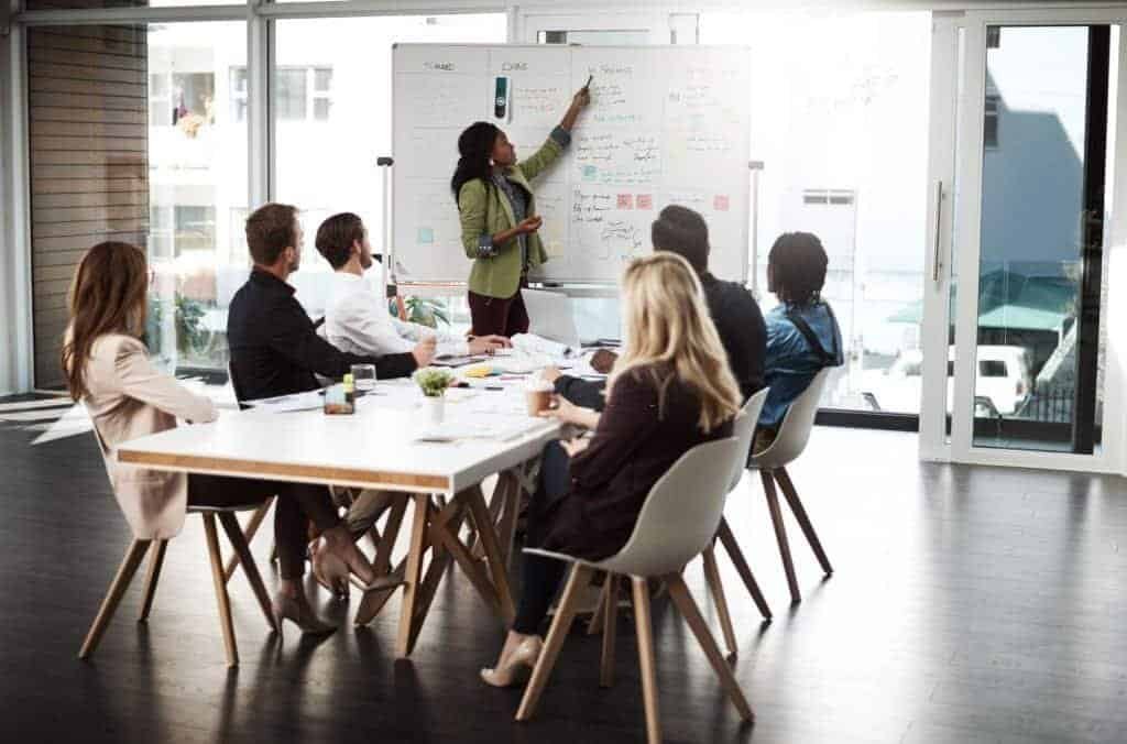 A group of business professionals seated around a table in a modern meeting room, listening to a woman presenting information on a whiteboard. The image reflects teamwork and collaborative discussions during a business strategy session.